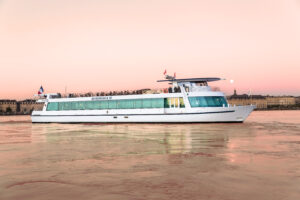 Le Burdigala 2 sous un ciel rose en soirée avec la Place de la Bourse en arrière-plan Le Burdigala 2 sous un ciel rose en soirée, arborant le drapeau français. Le bateau est visible en entier, avec en arrière-plan la Place de la Bourse de Bordeaux